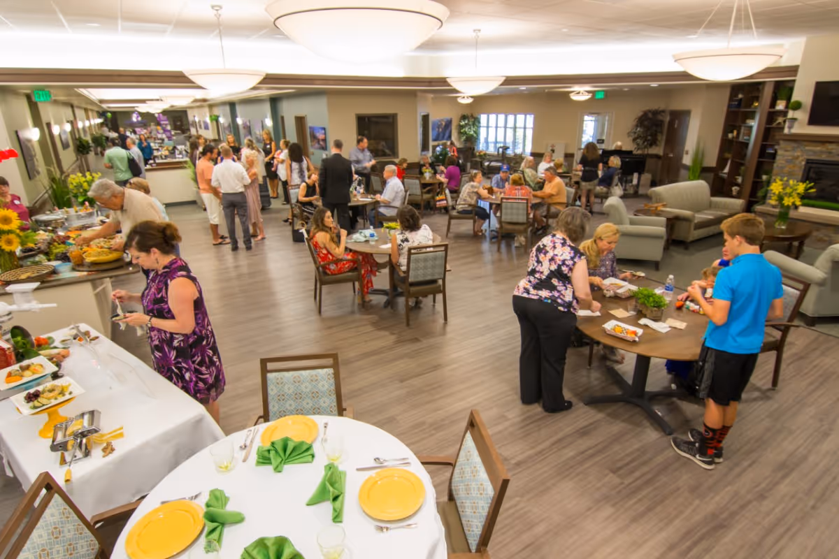 A large dining and common area in a senior living facility with multiple people socializing and eating. Several round tables are set with yellow plates and green napkins. There is a buffet table with food on the left side. The room has modern lighting, wood flooring, and comfortable seating including sofas near a fireplace.