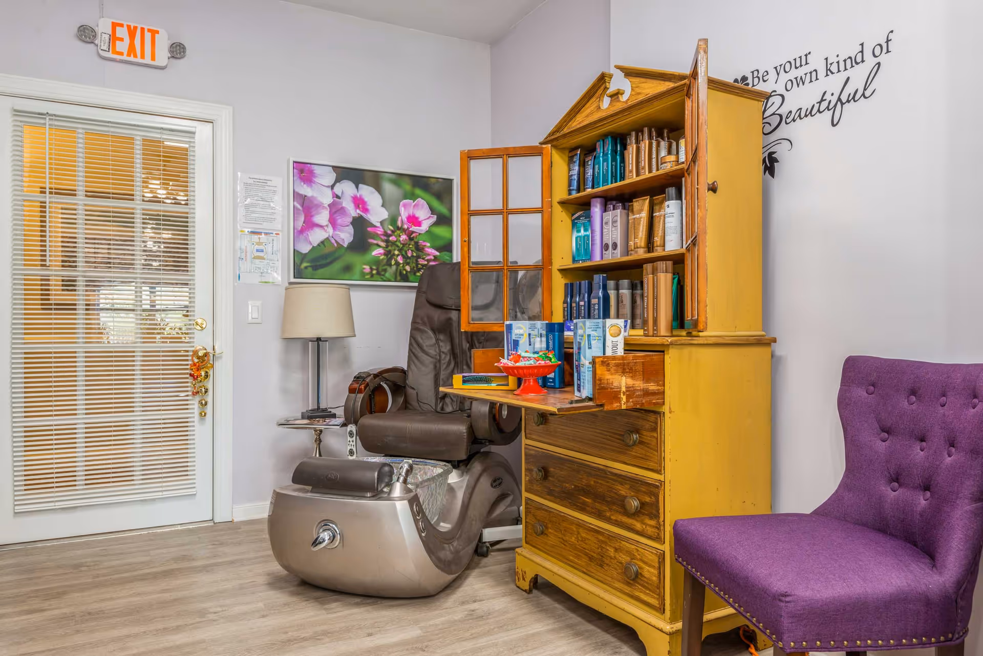 Interior view of a salon or spa area with a pedicure chair, a yellow wooden cabinet filled with hair and beauty products, a purple upholstered chair, a floor lamp, a floral wall picture, and an exit door with blinds.