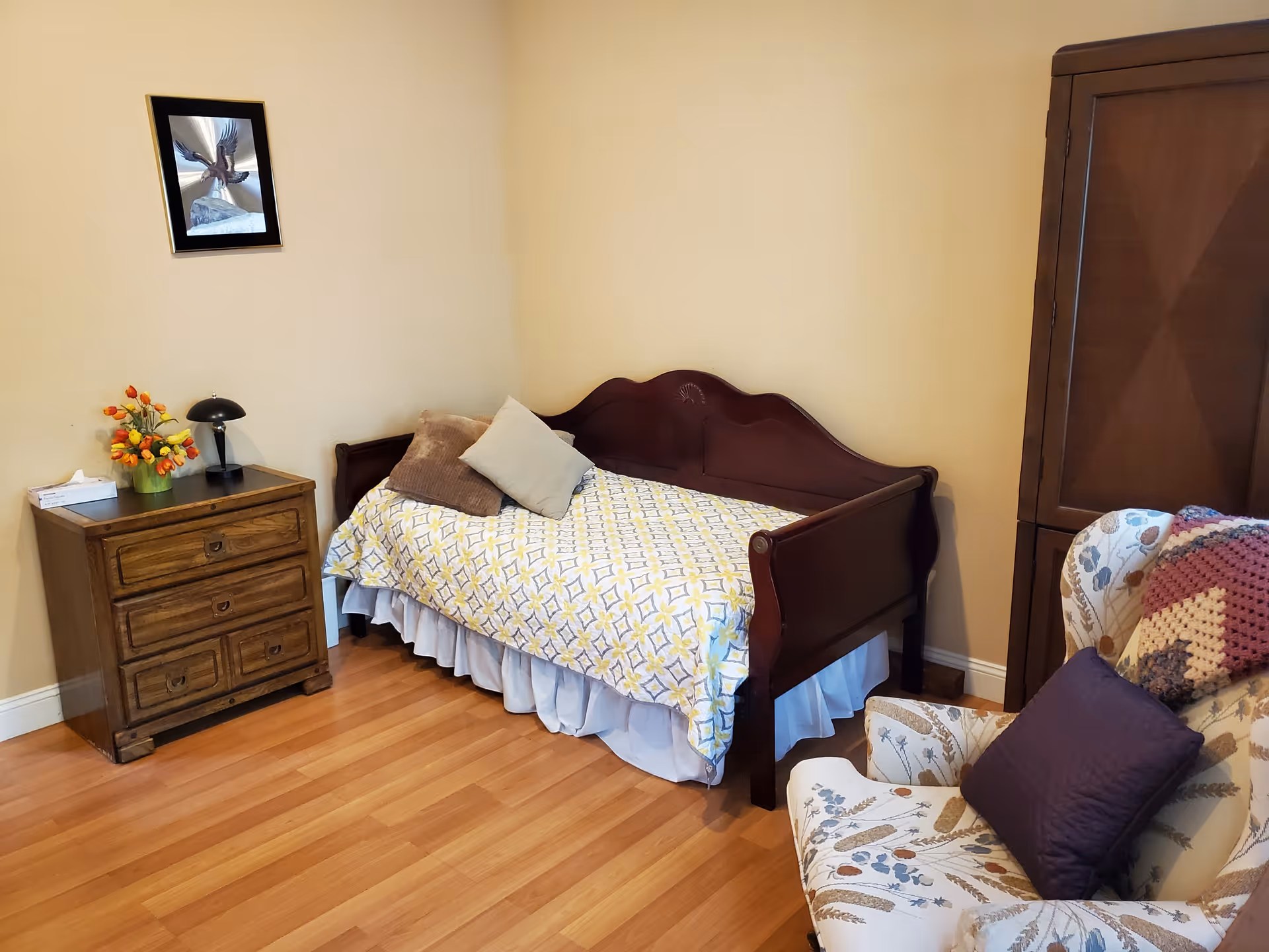 A cozy bedroom with a wooden daybed covered with a patterned yellow and white bedspread and two pillows. Next to the bed is a wooden nightstand with three drawers, a small black lamp, a tissue box, and a vase with orange and yellow flowers. A floral-patterned armchair with a purple pillow and a crocheted blanket is positioned near a wooden armoire. The walls are painted beige, and there is a framed picture hanging above the nightstand.