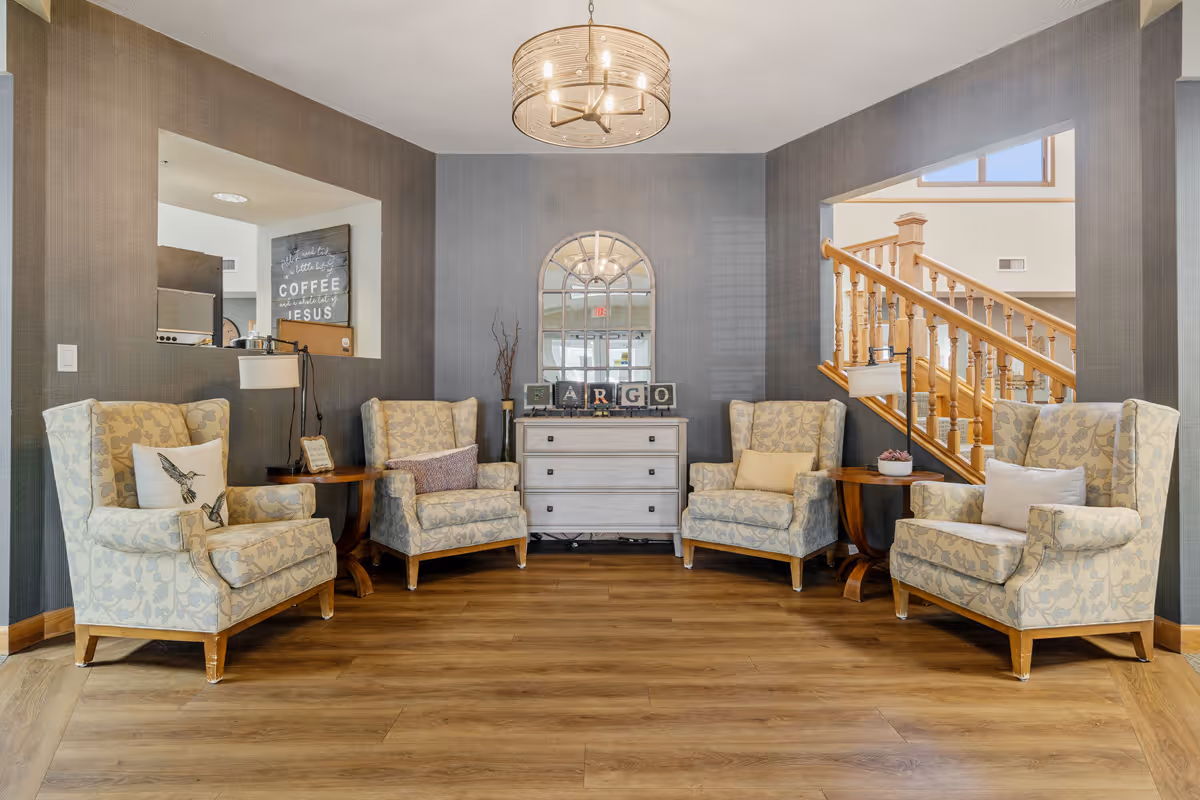 A cozy seating area in a senior living community featuring four patterned armchairs arranged around two wooden side tables. A white chest of drawers with decorative items and a large arched mirror is centered against a gray wall. A wooden staircase with spindles is visible on the right side, and a modern chandelier hangs from the ceiling. The floor is wood, and the space has a warm, inviting atmosphere.