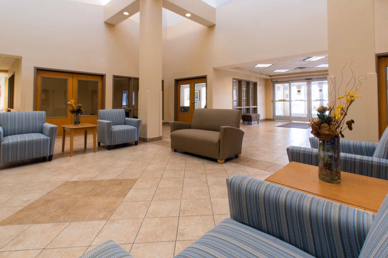 A spacious and well-lit common area with tiled floors, beige walls, and high ceilings. The room features several upholstered chairs in blue stripes and a brown loveseat arranged around wooden tables with floral centerpieces. There are large windows and glass doors in the background allowing natural light to enter.