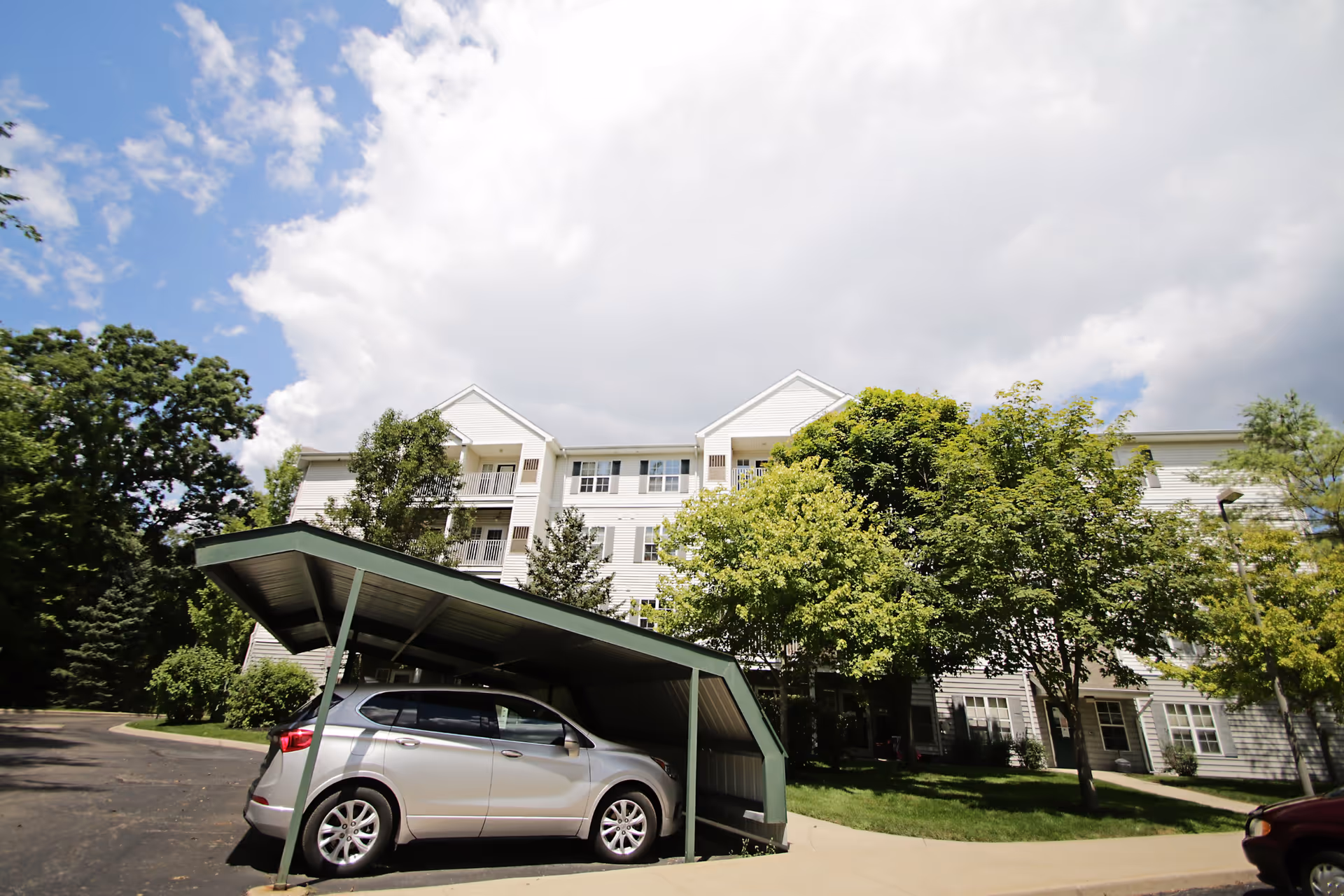 Exterior view of a multi-story senior living facility building with white siding, surrounded by green trees and a partly cloudy sky. In the foreground, there is a silver car parked under a green metal carport.