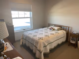 A simple bedroom with a bed covered by a quilted bedspread, a window, dresser and nightstand.