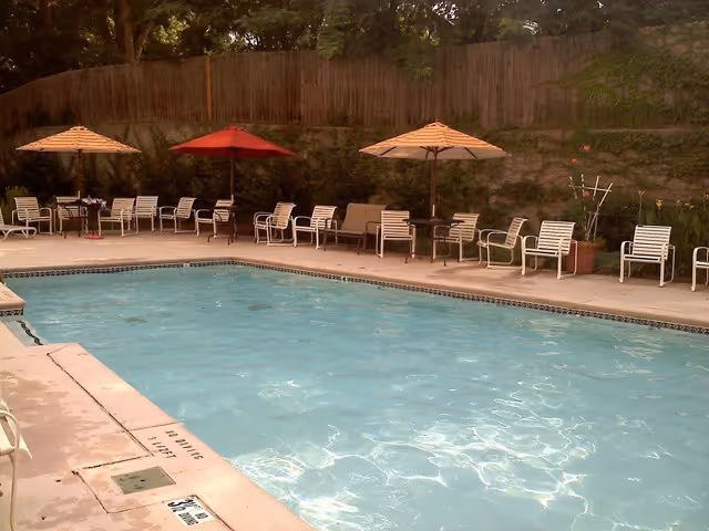 Outdoor swimming pool with lounge chairs and umbrellas along the far pool deck beside a wooden fence.