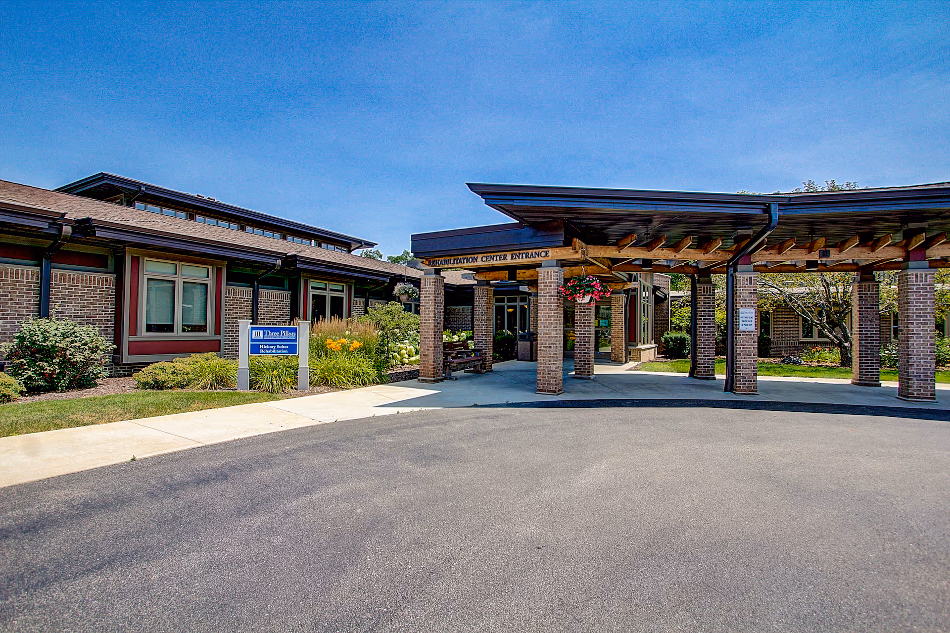 Front entrance of a brick senior living building with a covered drop-off area, columns, hanging flower baskets, and landscaped shrubs.