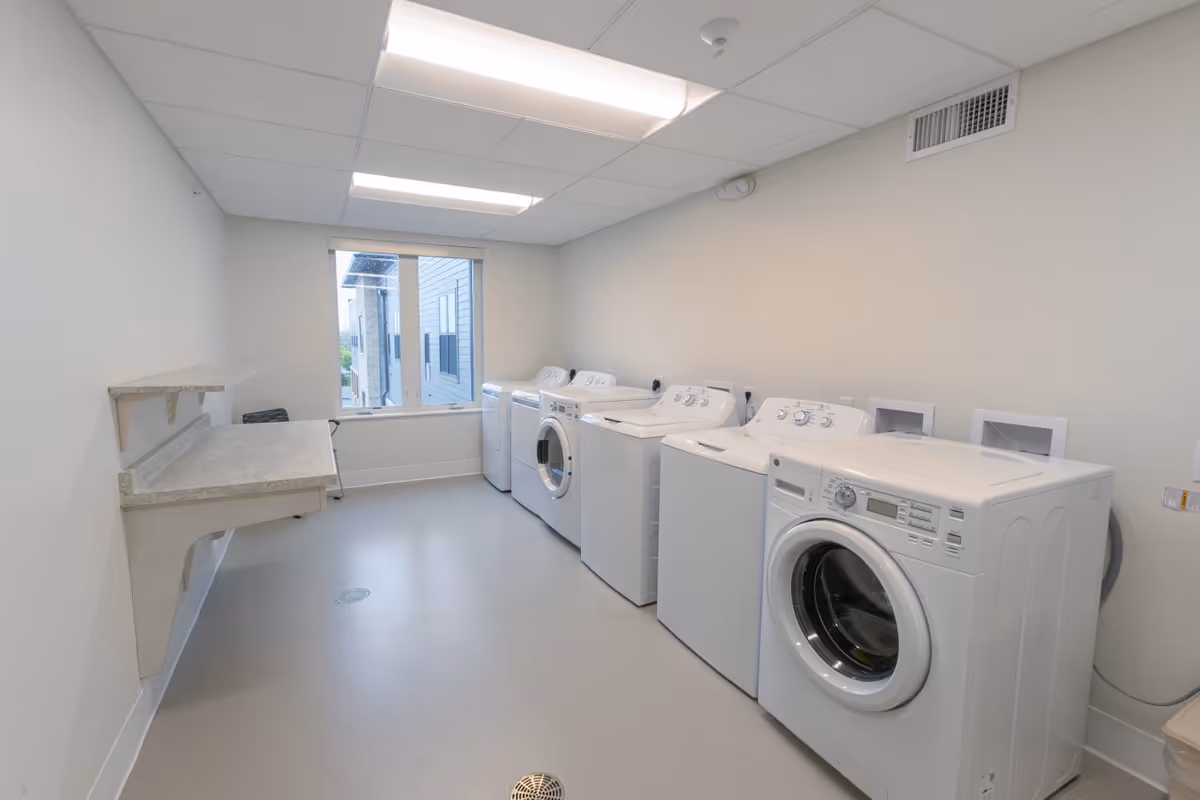 A clean and bright laundry room with multiple white washing machines and dryers lined up against the right wall. There is a folding counter attached to the left wall and a window at the far end letting in natural light. The room has a light-colored floor and ceiling with fluorescent lighting.