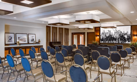Well-lit meeting room with rows of blue-upholstered chairs facing a large screen and framed artwork on the side walls.