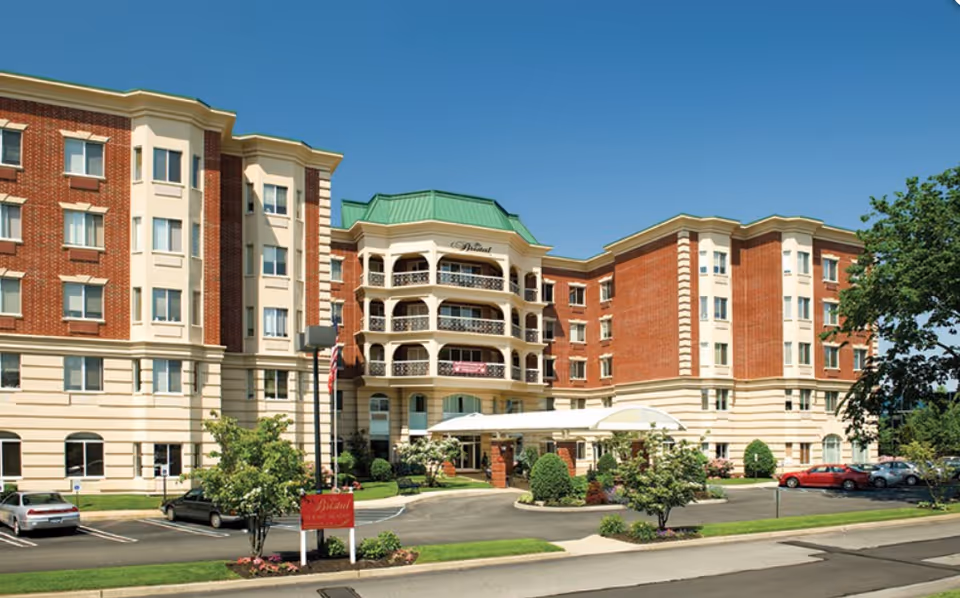 Front exterior of a large multi-story brick and beige assisted living building with balconies, a covered entrance, landscaping, and parked cars.