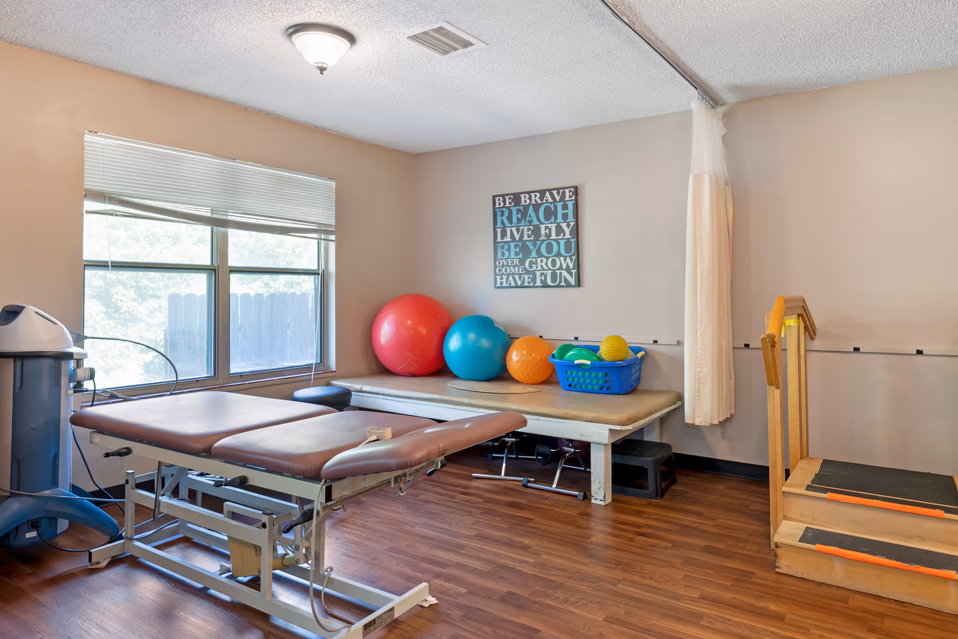 Physical therapy room with an adjustable treatment table, exercise balls, a basket with therapy equipment, and a small set of wooden stairs with handrails. The room has a window with blinds, wood flooring, and a motivational wall sign.