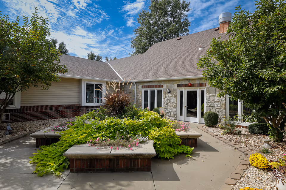Entrance courtyard of an assisted living building featuring raised flower beds, shrubs, and double glass doors.