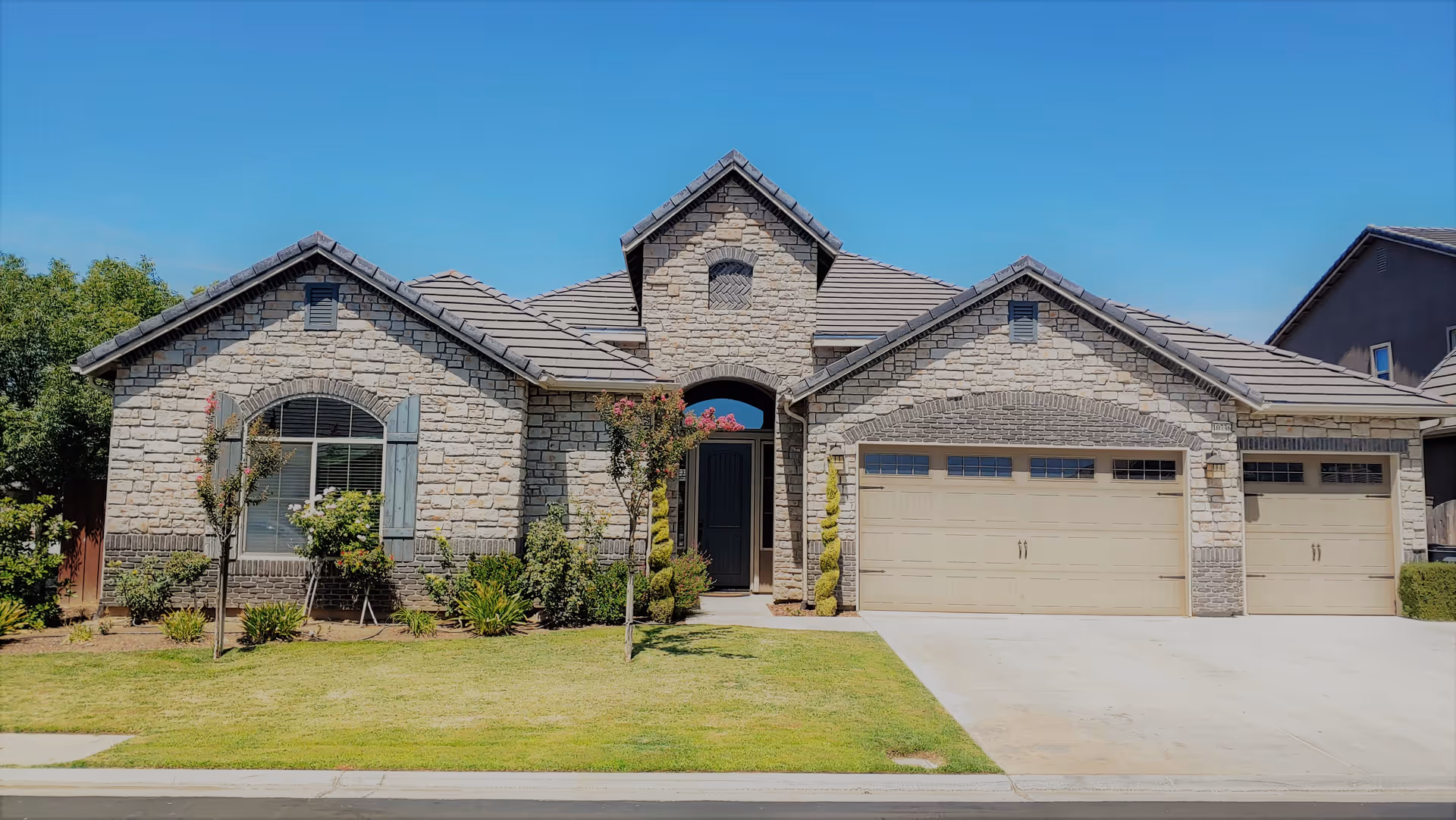 Front exterior view of a single-story residential building with stone facade, three garage doors, a central entrance with an arched window above, and a well-maintained lawn with small trees and shrubs under a clear blue sky.