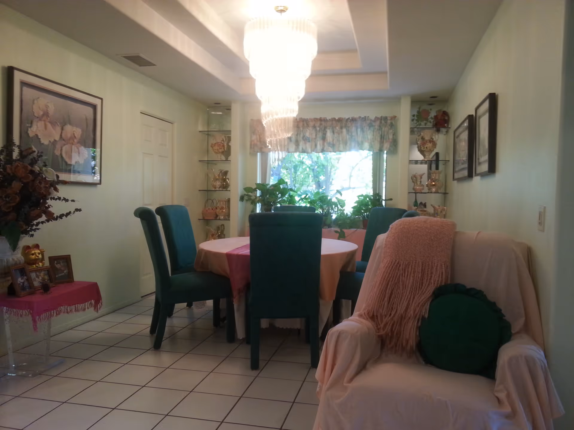 A dining room with a round table covered with a tablecloth and surrounded by six green upholstered chairs. There is a large window with floral curtains and several potted plants on the windowsill. A white armchair with a pink throw blanket and a green round pillow is in the foreground. The room has tiled flooring, light green walls, framed pictures, and glass shelves displaying decorative items. A chandelier hangs from the ceiling above the table.