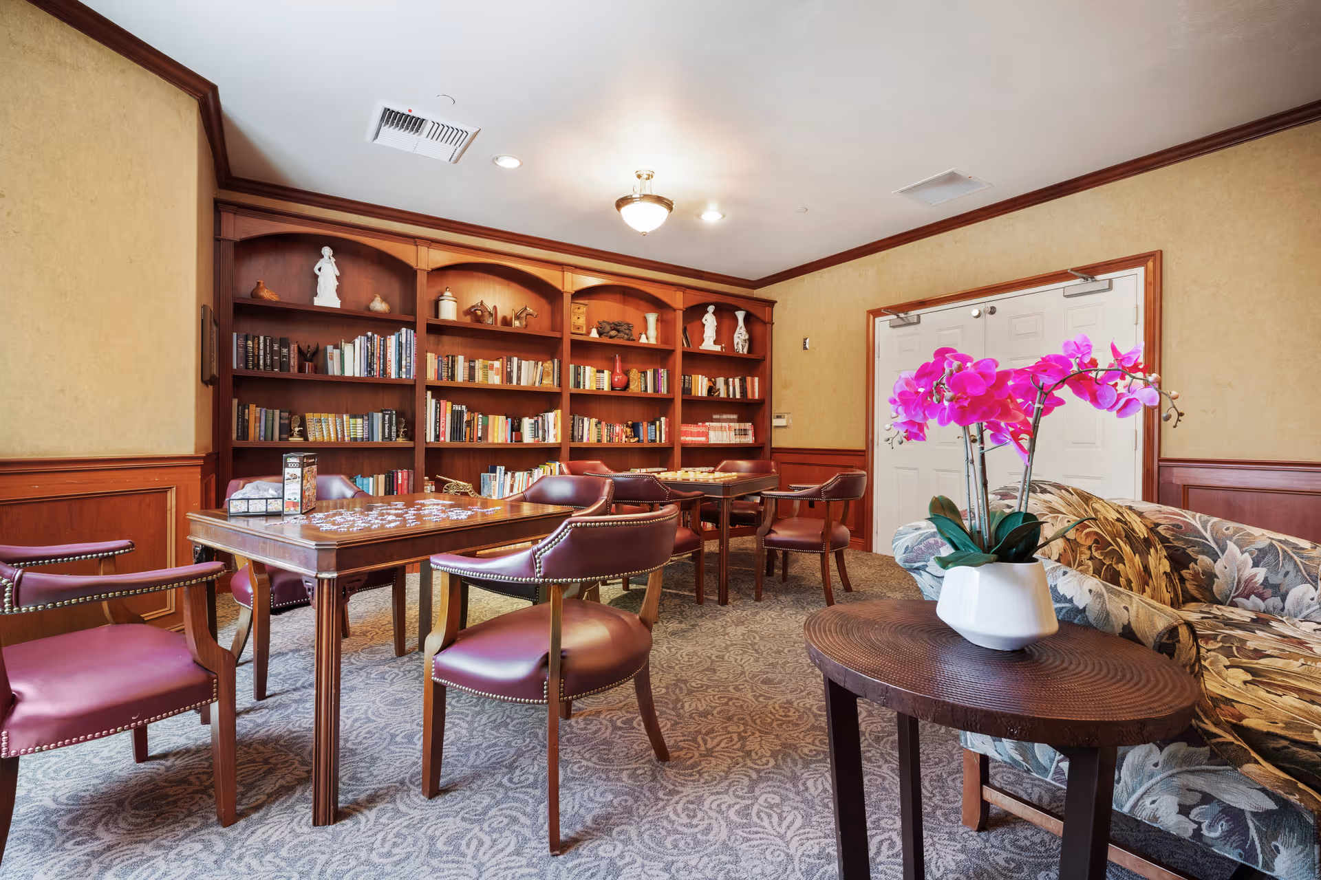 A cozy room with wooden bookshelves filled with books and decorative items, several wooden tables with chairs arranged around them, a floral patterned sofa, and a small round wooden table with a white vase holding vibrant pink orchids.