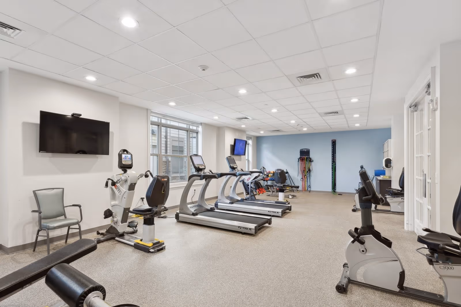A bright and spacious fitness room in a senior living facility featuring several exercise machines including treadmills, stationary bikes, and a rowing machine. The room has a light-colored ceiling with recessed lighting, large windows letting in natural light, and a wall-mounted TV. There are also chairs and various fitness equipment along the walls.