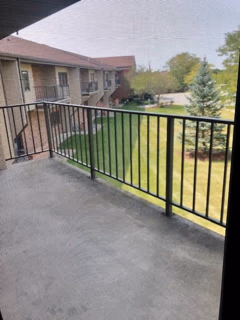 View from a balcony with a black metal railing overlooking a grassy area with trees and a two-story building with balconies in the background.