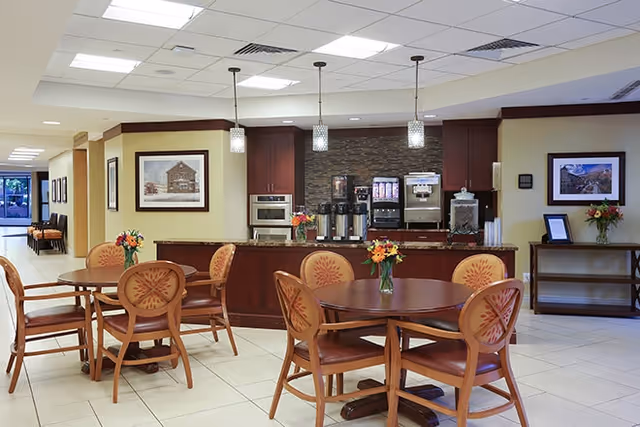 A dining area with round wooden tables and chairs featuring floral upholstery. Each table has a small vase with colorful flowers. In the background, there is a counter with coffee and beverage dispensers, cabinets, and framed artwork on the walls. The space is well-lit with ceiling lights and pendant lights above the counter.