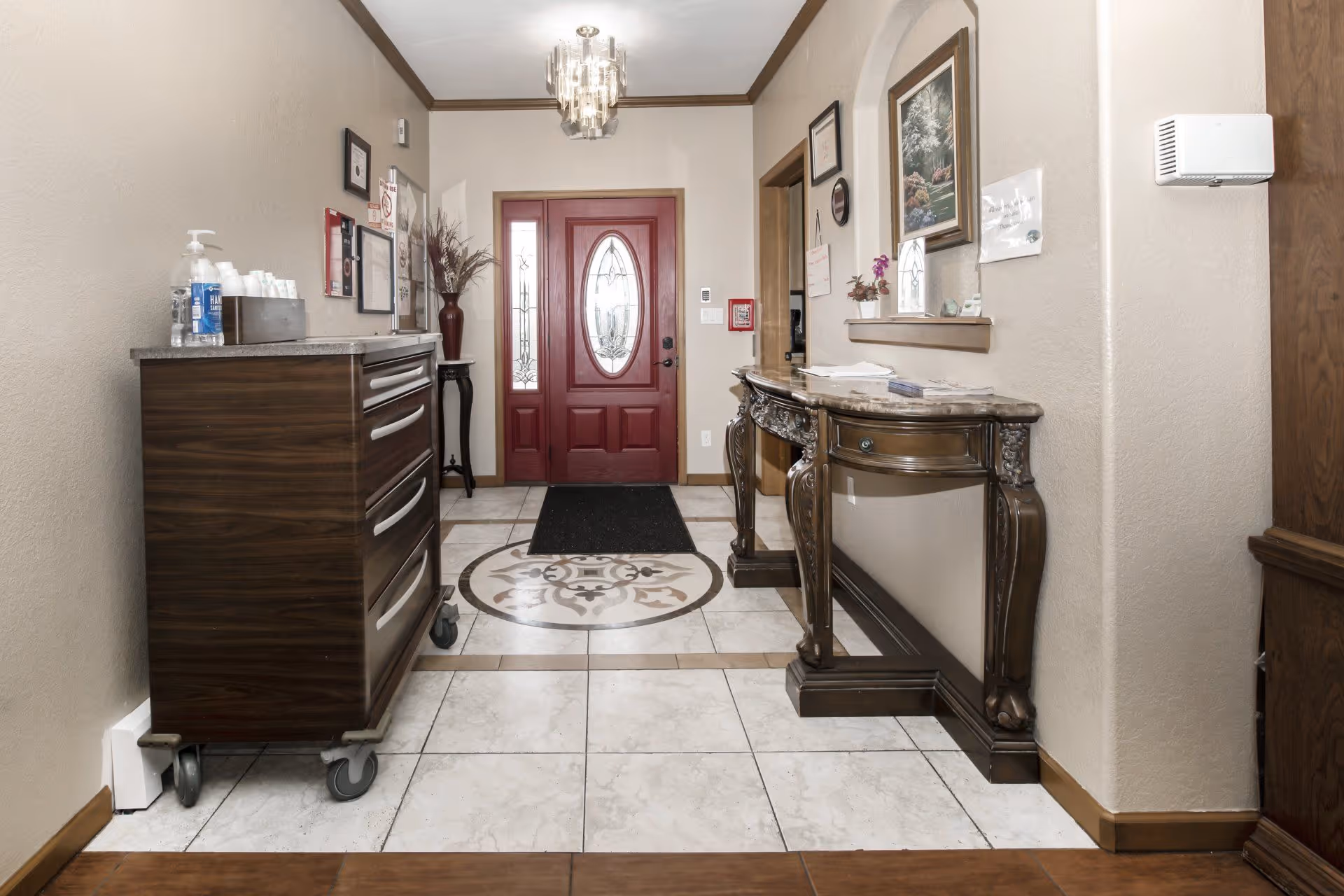 Interior hallway with a decorative red front door featuring glass panels. The floor is tiled with a circular ornamental design in the center. On the left side, there is a dark wooden cabinet on wheels with hand sanitizer and cups on top. On the right side, there is an ornate wooden console table with a marble top, a framed painting above it, and some papers and a small plant on the table. The walls are beige with wooden trim, and a chandelier hangs from the ceiling.