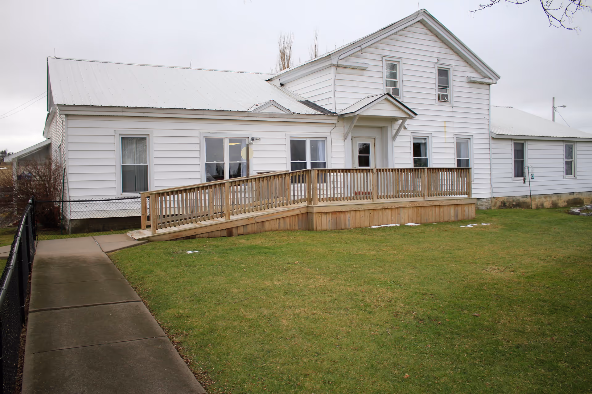 Exterior view of a white two-story building with a wooden wheelchair ramp leading to the entrance, surrounded by a green lawn and a concrete walkway on the left side.