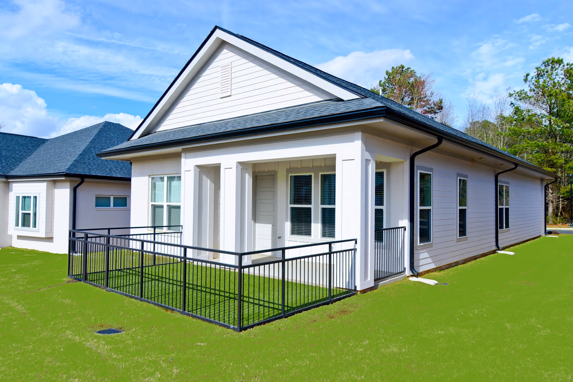 Exterior view of a single-story white building with a black roof and multiple windows. The building has a small fenced porch area with a black metal railing and green grass surrounding it. Another similar building is partially visible in the background under a blue sky with some clouds.