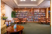 A cozy library room in a retirement community featuring wooden bookshelves filled with books along the back wall, several wooden tables with chairs arranged for reading or studying, a green carpeted floor, and a small round wooden coffee table with a decorative item in the foreground. The room is softly lit with ceiling lights and a table lamp on the left side near a beige sofa.