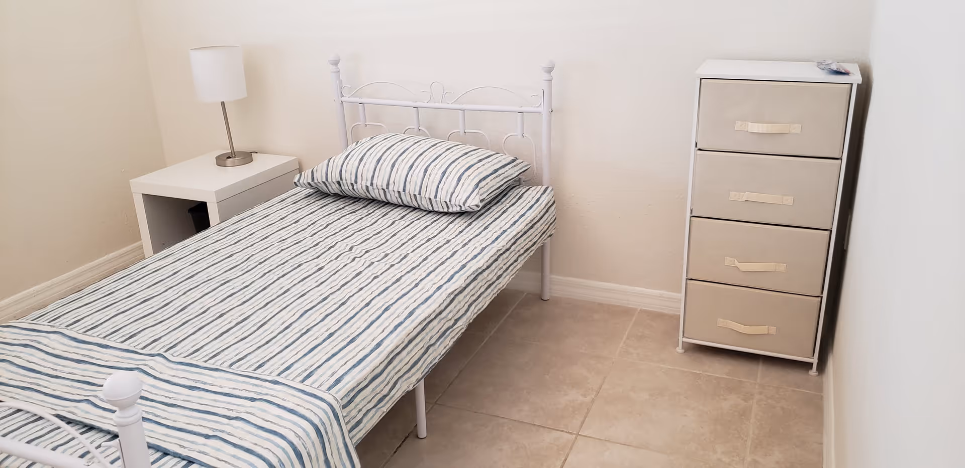 A small bedroom with a single white metal bed frame, a striped bed sheet and pillow, a white nightstand with a lamp, and a beige four-drawer fabric dresser against a white wall.