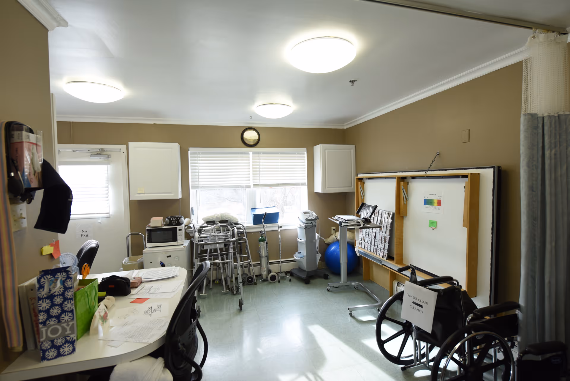 Interior therapy/equipment room with wheelchairs, walkers, a table with paperwork and a whiteboard.