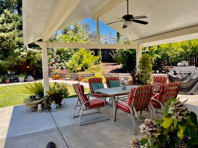 Covered outdoor patio area with a ceiling fan, a table surrounded by eight red and white striped cushioned chairs, and various potted plants. In the background, there is a garden with green shrubs, trees, and a wooden fence under a clear blue sky.