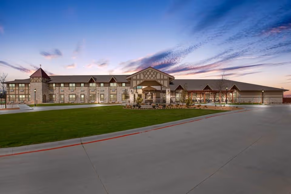 Exterior view of a large senior living facility building named The Villages of Windcrest during sunset, featuring a well-maintained lawn, a driveway, and architectural details including stone and wood accents.