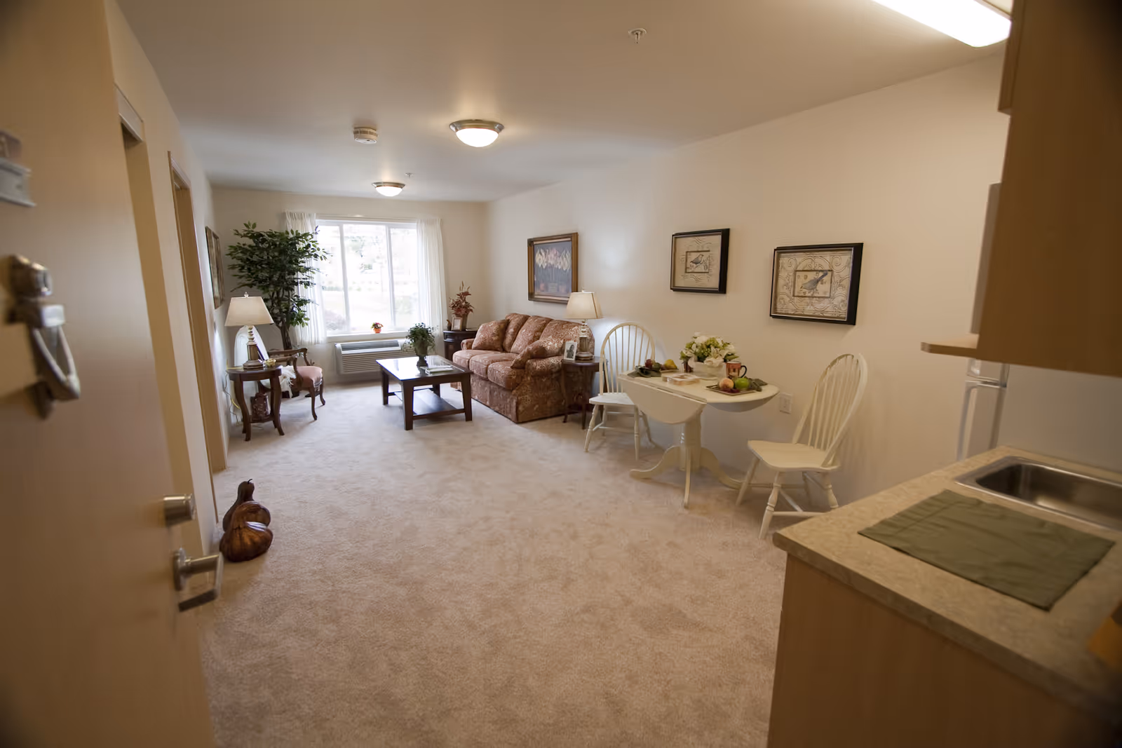 Interior view of a senior living apartment at Amber Park Senior Living showing a living room area with a patterned sofa, coffee table, side tables with lamps, and framed artwork on the walls. There is a small dining table with two white chairs set with fruit and flowers. A window with white curtains lets in natural light. The foreground includes a kitchenette with a sink and countertop.