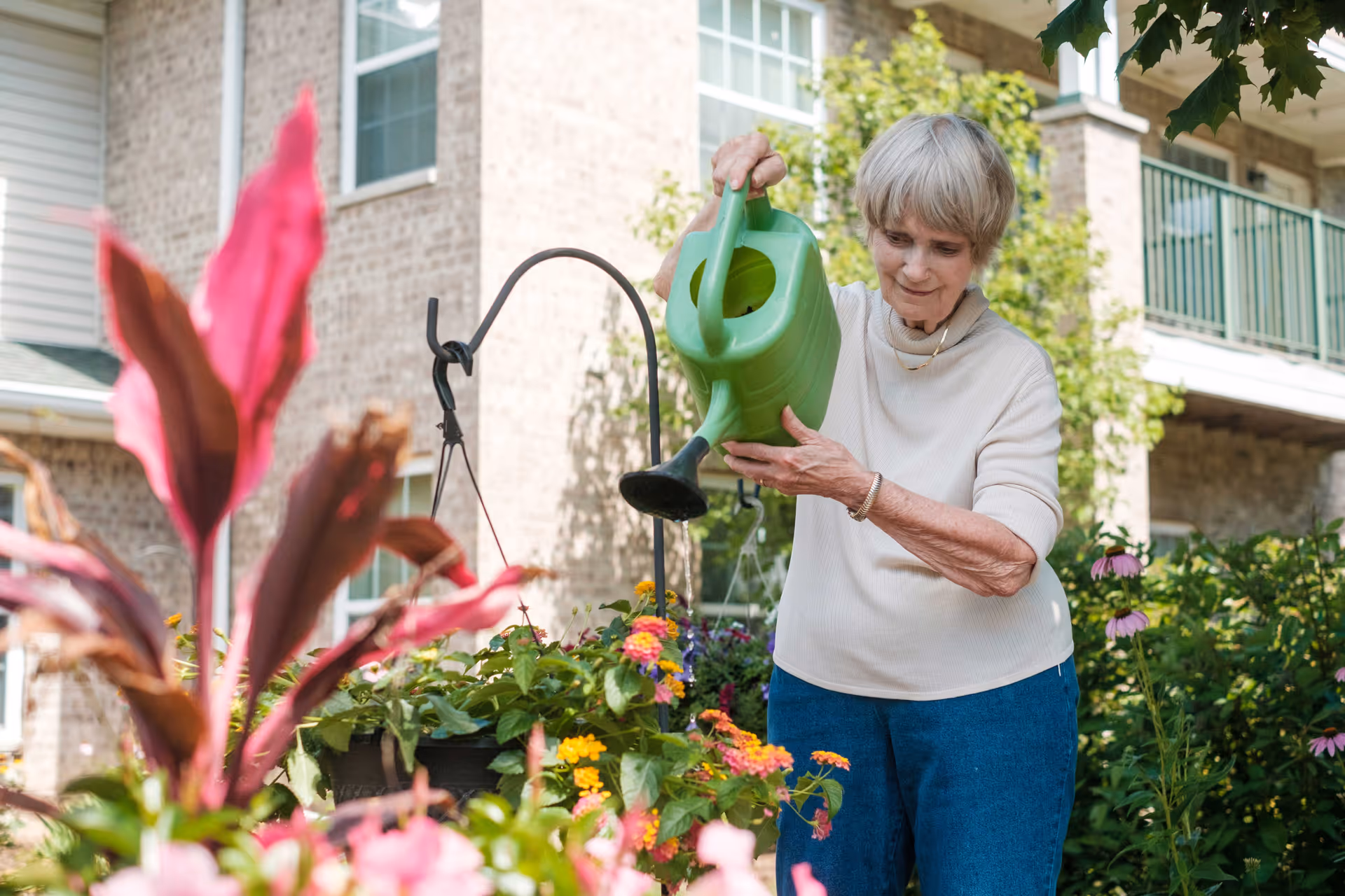 An elderly woman watering colorful flowers in a garden outside a brick building with balconies, enjoying outdoor gardening.