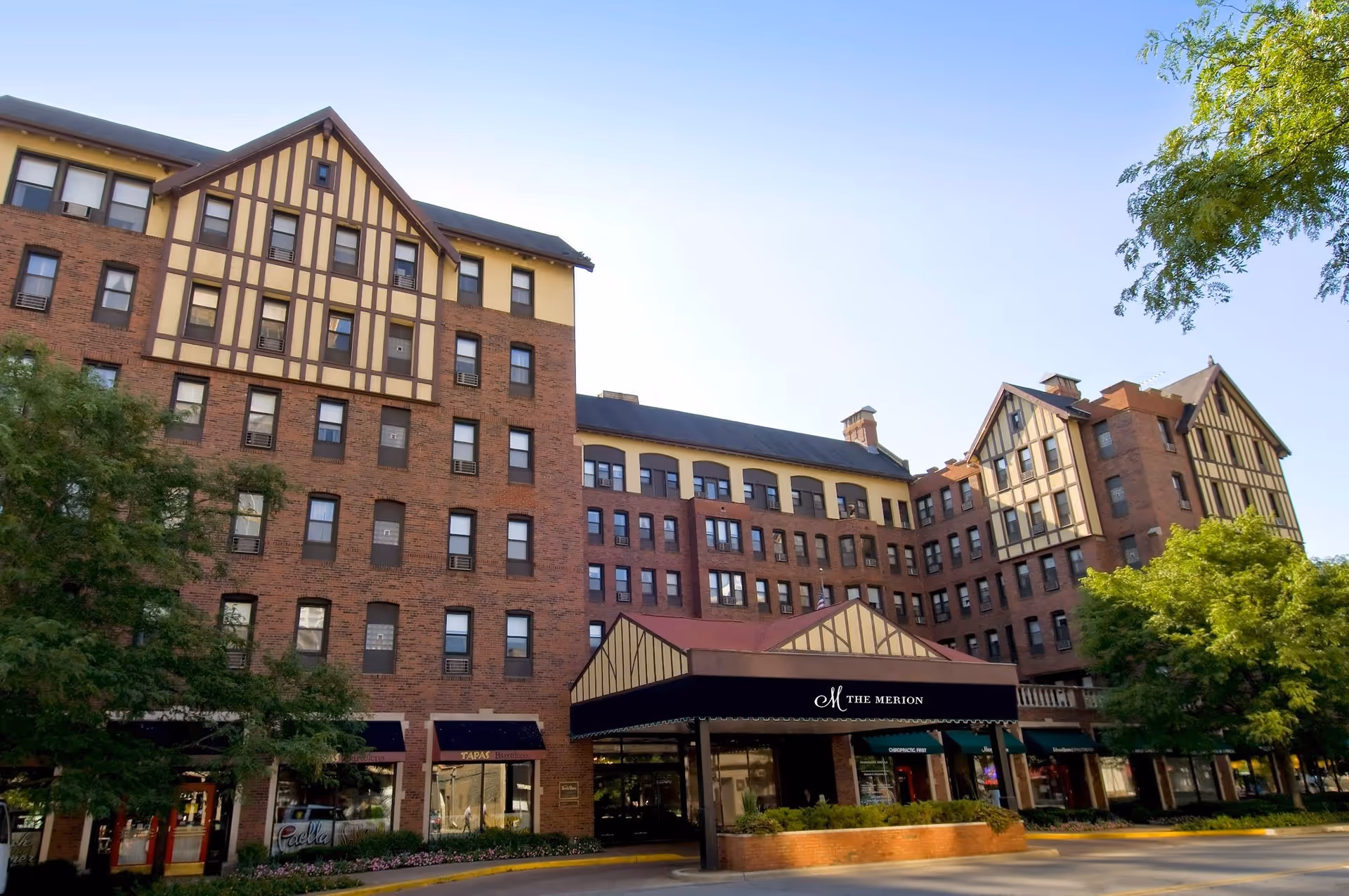 Exterior view of The Merion, a multi-story brick building with Tudor-style architectural accents, large windows, and a covered entrance. Trees and greenery surround the building under a clear blue sky.