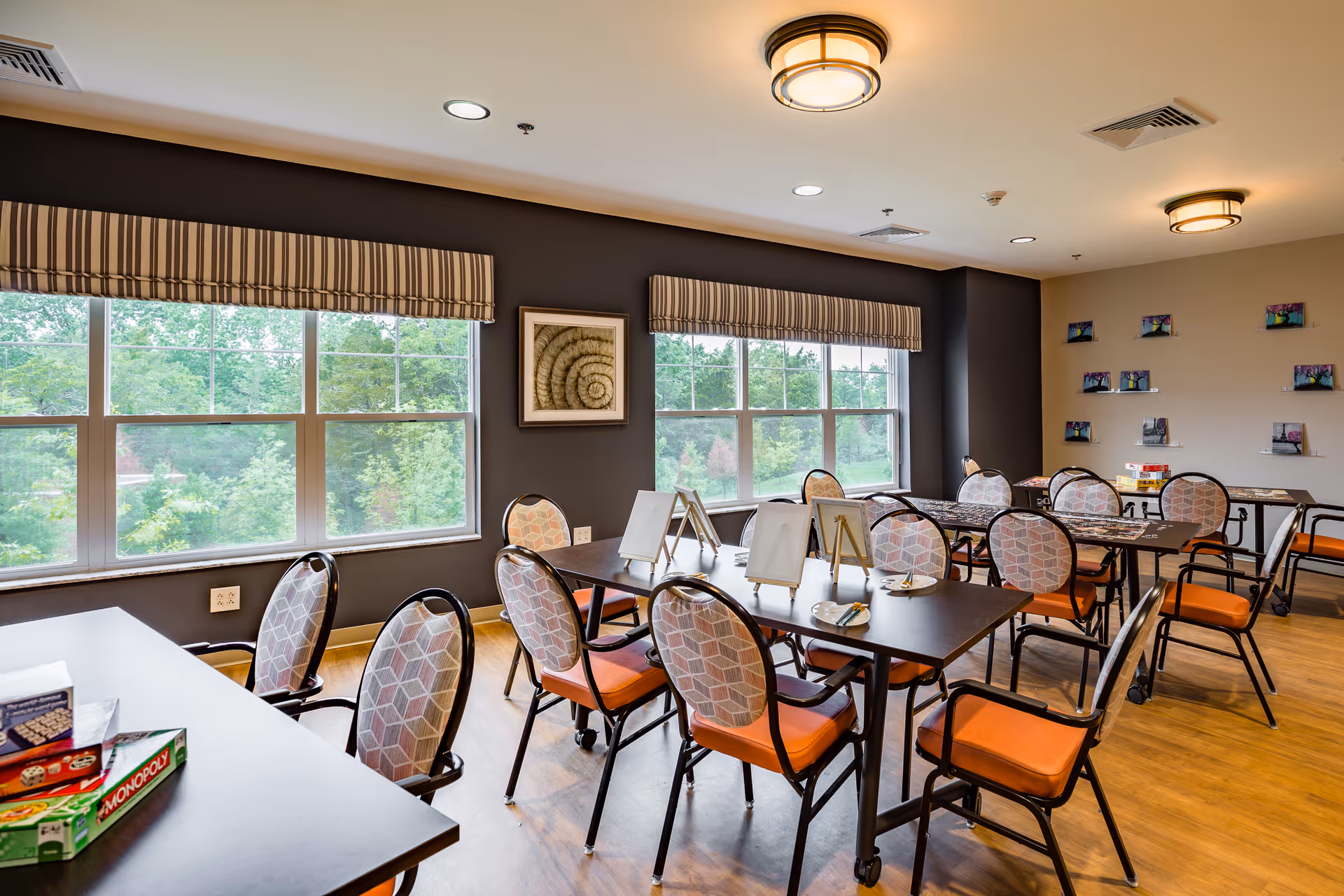 A bright activity room with multiple tables and chairs arranged for group activities. The chairs have patterned backs and orange seats. There are large windows with striped valances letting in natural light, and framed artwork on the walls. Board games like Monopoly are visible on one of the tables.