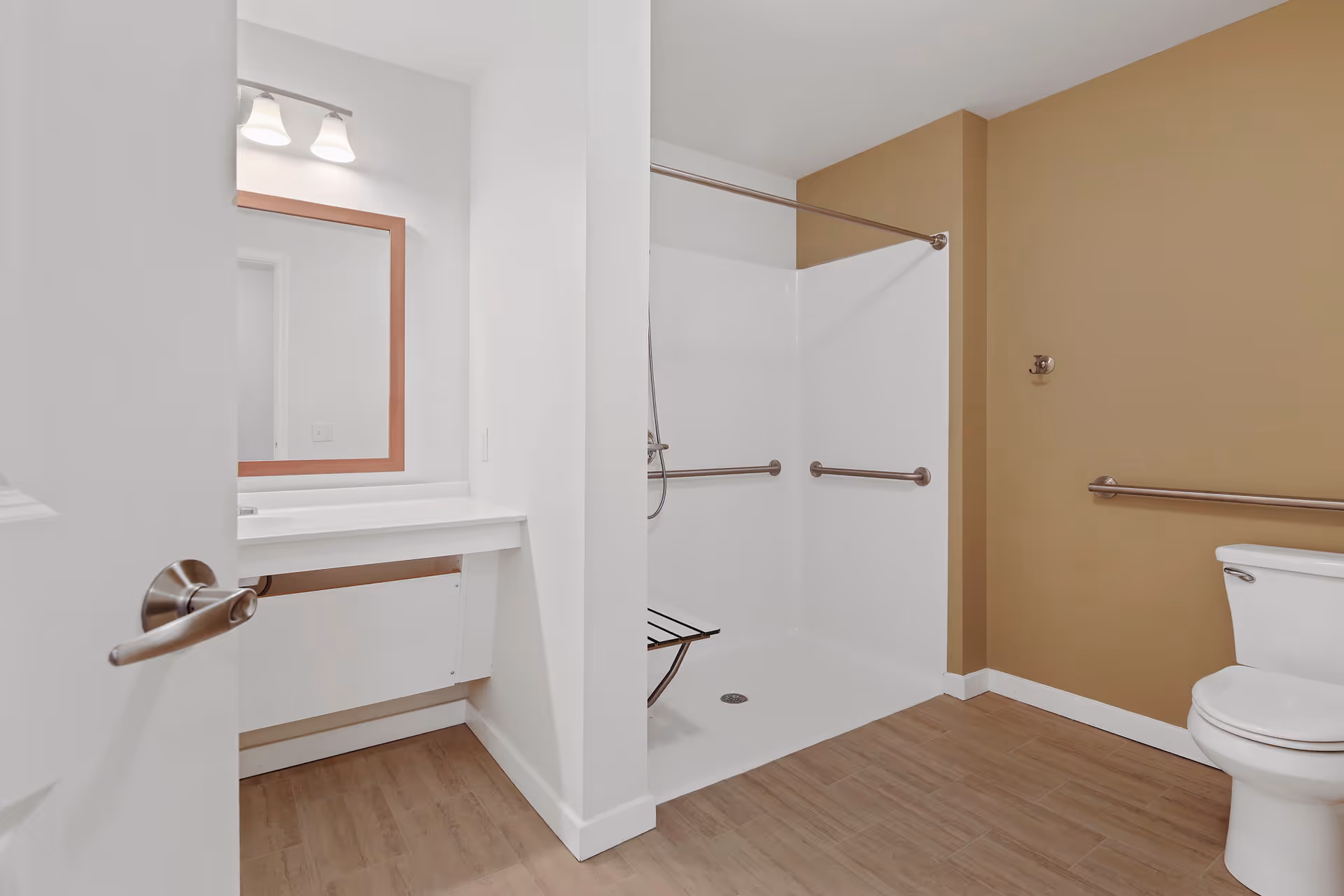 A spacious bathroom with a walk-in shower featuring grab bars and a foldable shower seat. The bathroom has a toilet with a grab bar on the wall beside it, a vanity with a mirror and two light fixtures above it. The floor has wood-like tiles and the walls are painted white and beige.