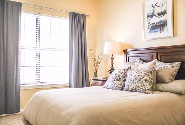 A bright bedroom with a large window covered by gray curtains, a bed with a wooden headboard, beige bedding, and several decorative pillows. A nightstand with a lamp and a vase with branches is next to the bed, and a framed abstract painting hangs on the wall above the headboard.