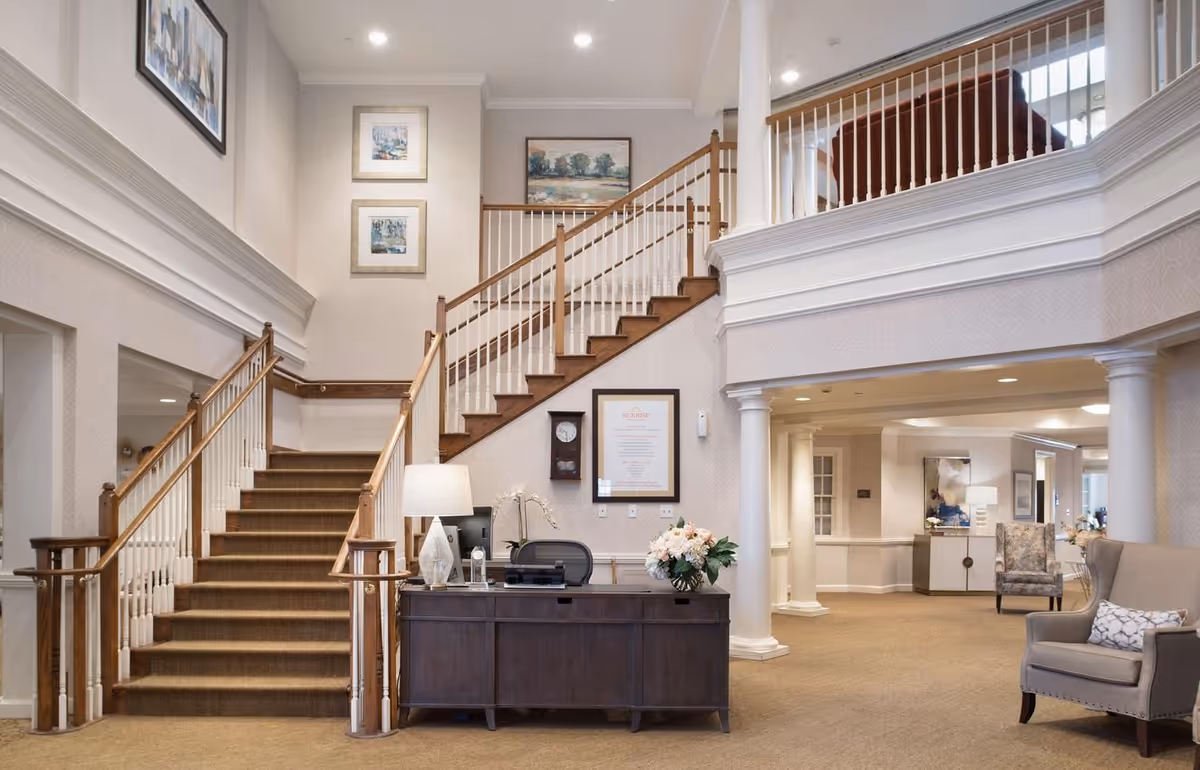 Bright, elegant senior living lobby with a central staircase, reception desk, and seating area.
