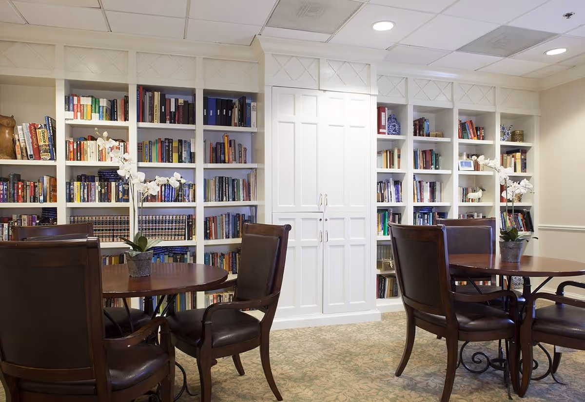 A cozy library room with built-in white bookshelves filled with books and decorative items. There are two round wooden tables, each surrounded by four dark brown leather chairs. Two potted white orchid plants are placed on the tables. The room has a carpeted floor and a white paneled ceiling with recessed lighting.