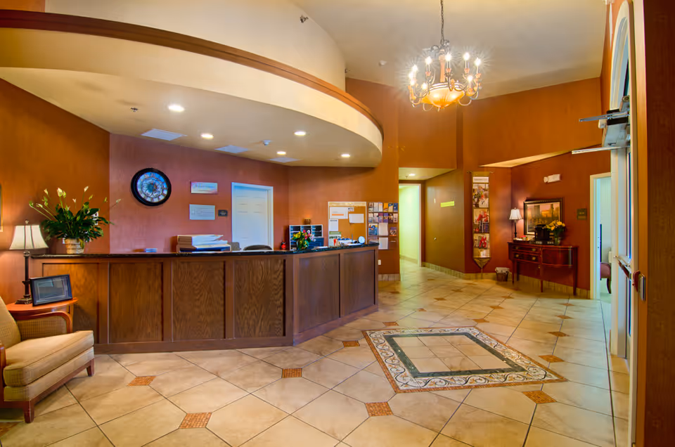 Reception area of Parkside Manor Assisted Living and Memory Care featuring a wooden front desk with office supplies, a clock on the wall, a comfortable chair, decorative plants, a chandelier, and a tiled floor with a decorative square pattern in the center.