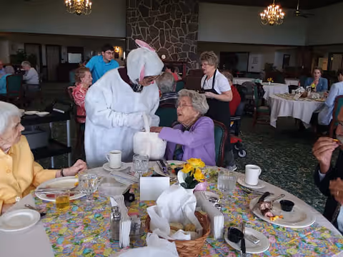 A group of elderly people seated around a dining table in a retirement community dining room. One person is dressed in a white bunny costume interacting with an elderly woman in a purple sweater. The table is set with plates, cups, and a floral tablecloth. Other residents and staff are visible in the background.