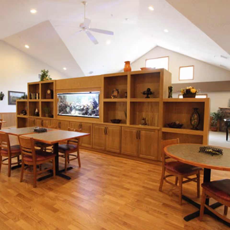 Interior view of a senior living facility common area with wooden flooring, tables and chairs, a large wooden shelving unit with decorative items and a built-in aquarium, and a ceiling fan above.