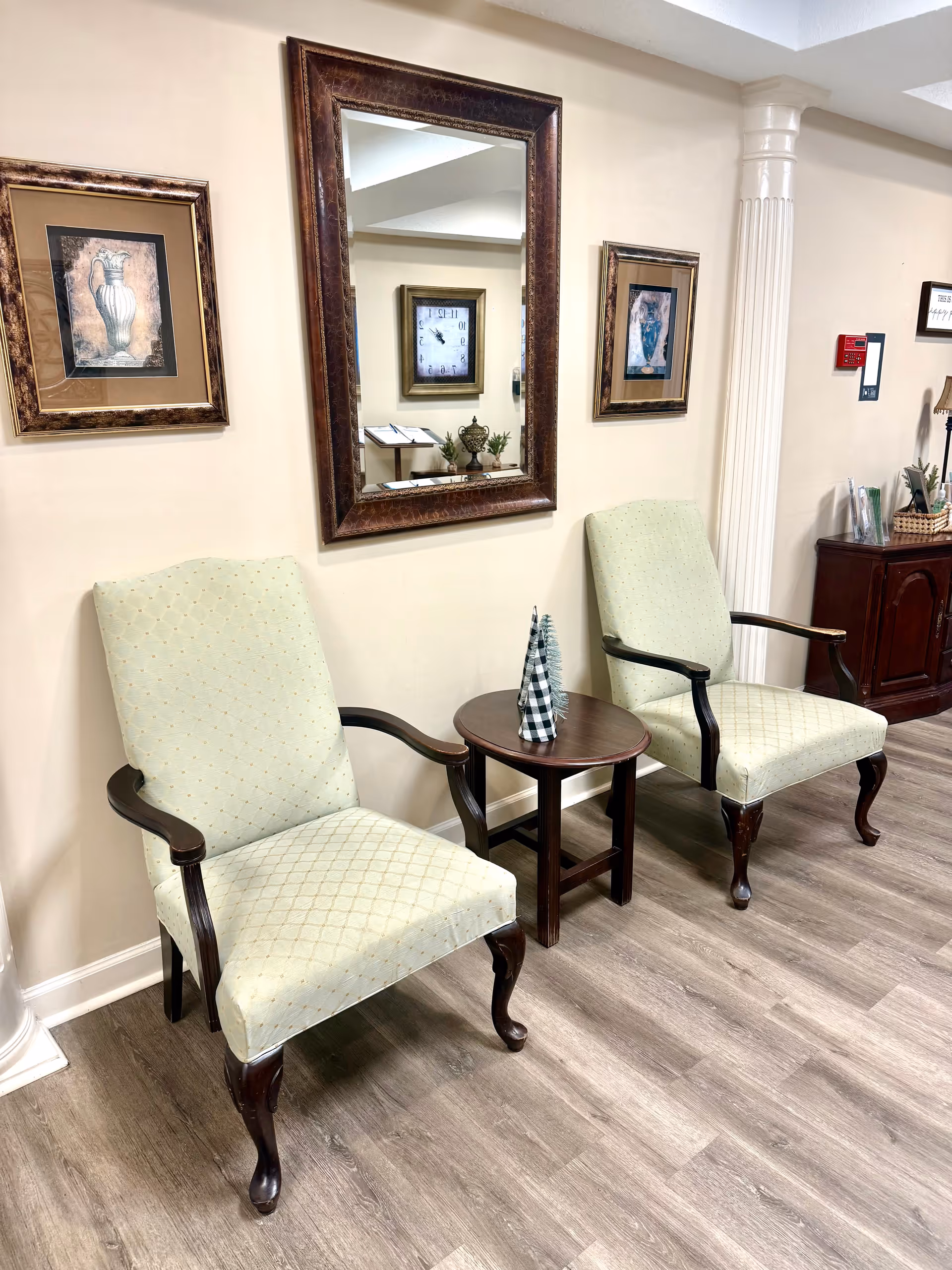Two upholstered armchairs with wooden legs and armrests are placed on either side of a small round wooden table with two decorative cone-shaped objects. Behind the chairs, a large framed mirror is mounted on a beige wall, flanked by two framed pictures of vases. The room has wood-look flooring and a white decorative column is visible on the right side.