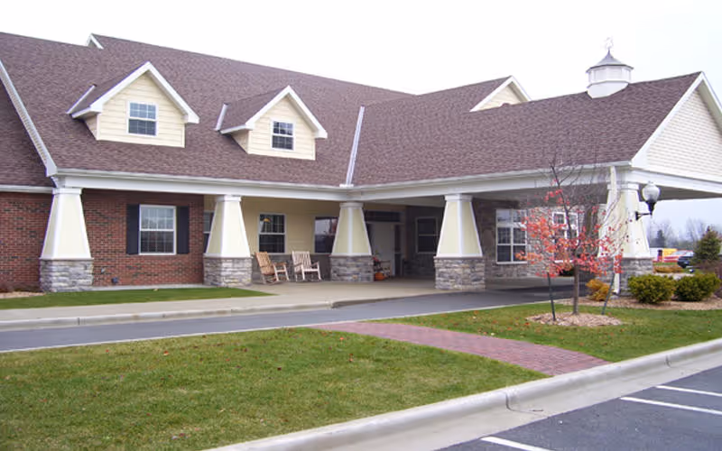 Exterior view of a single-story senior living facility building with a covered entrance supported by large columns. The building has a brown shingled roof with dormer windows, brick and stone accents on the walls, and a small landscaped area with a tree and bushes near the entrance. There are two rocking chairs on the porch area under the covered entrance.