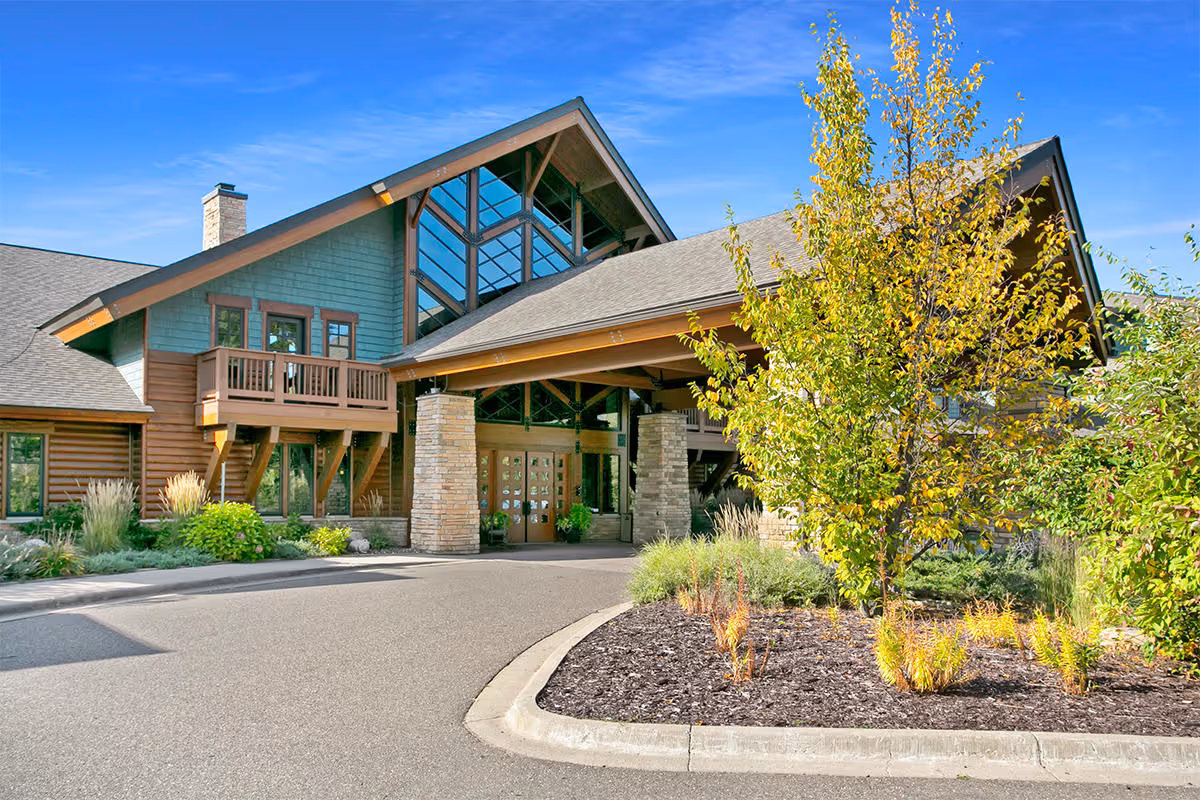 Exterior view of a senior living facility named Heartwood with a large covered entrance, stone pillars, wooden siding, and a landscaped area with trees and shrubs under a clear blue sky.