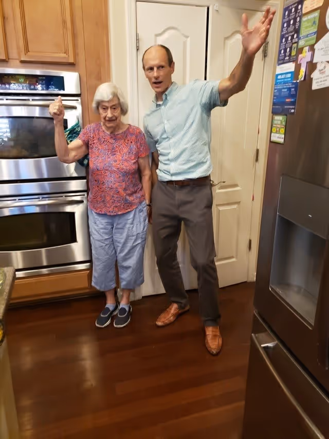 An elderly woman and a middle-aged man standing together in a kitchen. The woman is wearing a patterned pink shirt and light blue pants, and the man is wearing a light blue shirt and brown pants. They are both smiling and gesturing with their arms raised. The kitchen has wooden cabinets, a stainless steel double oven, and a stainless steel refrigerator with magnets and notes on it.