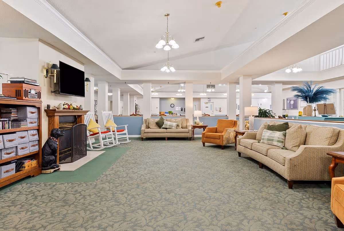 A spacious senior living facility common area with patterned carpet, beige sofas with green and plaid pillows, an orange armchair, white rocking chairs with yellow cushions, a wooden cabinet with storage bins, a fireplace with a black dog statue, and a wall-mounted flat-screen TV. The room is well-lit with ceiling lights and decorated with plants and artwork.