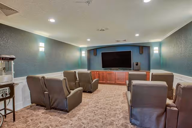 A cozy media room with six brown recliner chairs arranged in two rows facing a large flat-screen TV mounted on a blue accent wall. The room has beige carpet, white wainscoting on the walls, and soft overhead lighting. A popcorn machine is visible on the left side of the room.