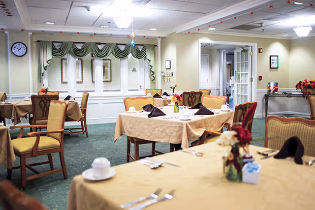 A dining room with several tables covered in beige tablecloths, each set with cups, saucers, silverware, and folded dark napkins. The room has green carpeting, cream-colored walls, and decorative curtains over a window. There are upholstered chairs around the tables and a clock on the wall.