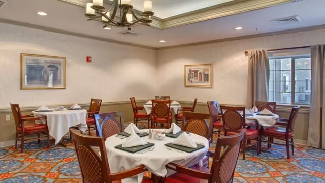 A dining room with round tables covered in white tablecloths, each set with folded napkins, menus, and silverware. The room has patterned carpet, framed artwork on the walls, a window with curtains, and a chandelier hanging from the ceiling.