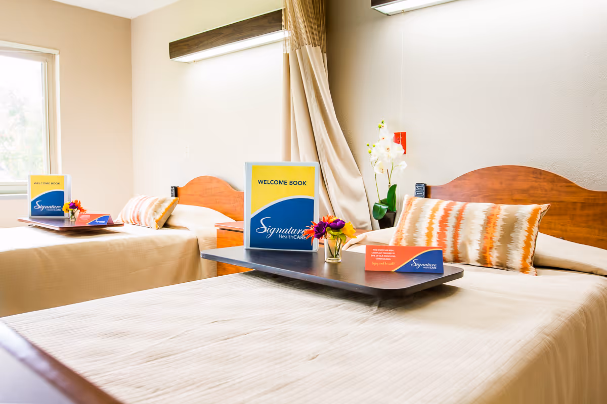 A bright and clean bedroom with two single beds, each with a wooden headboard, beige bedspreads, and decorative pillows. Each bed has a tray on it holding a welcome book, a small flower arrangement, and a card with the Signature HealthCARE logo. A window with beige curtains is visible in the background.
