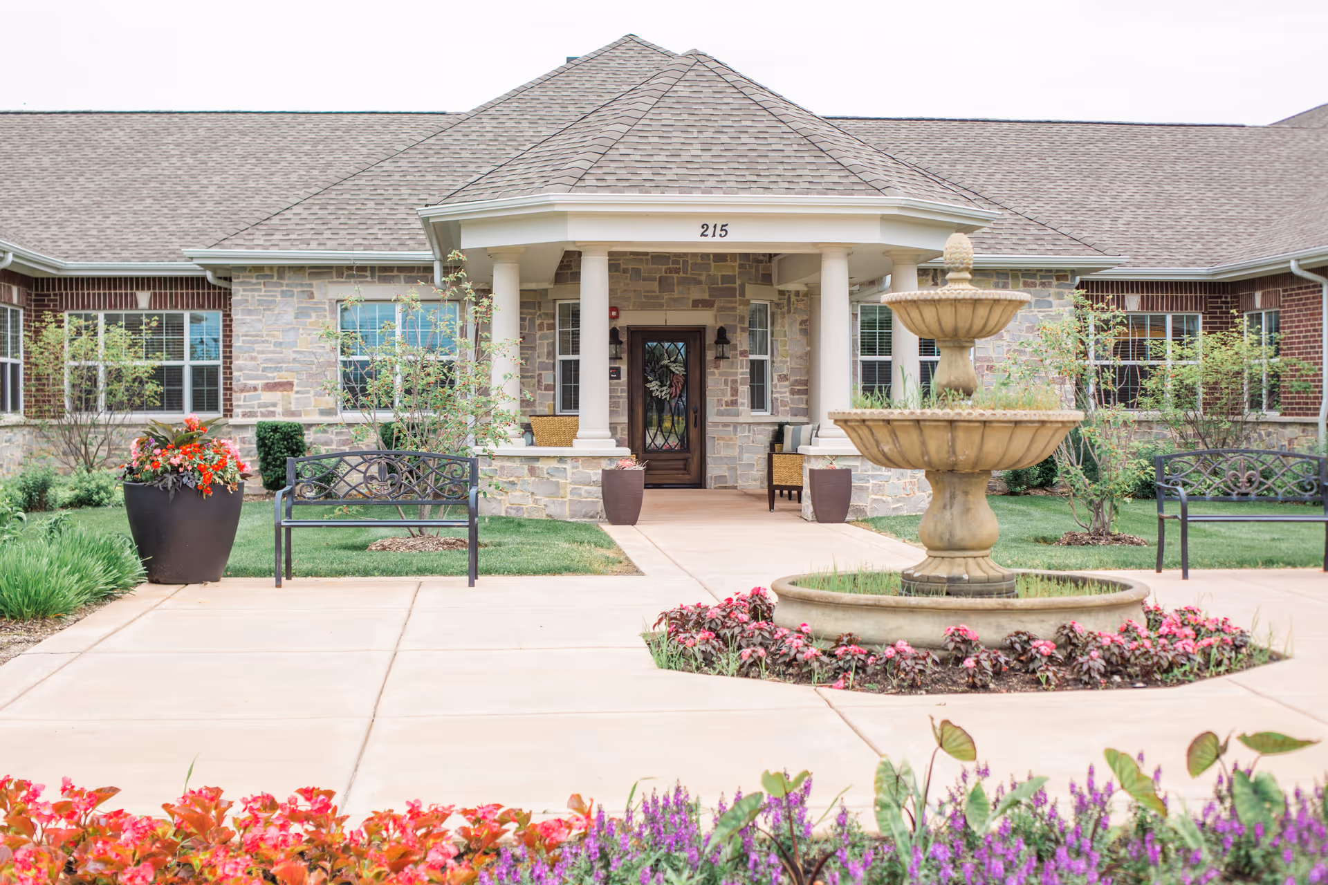 Front exterior view of a senior living facility with a stone facade, a covered entrance supported by white columns, a decorative glass door, and the number 215 above the entrance. The walkway leading to the entrance is flanked by benches, large planters with flowers, and a tiered stone fountain surrounded by colorful flower beds.