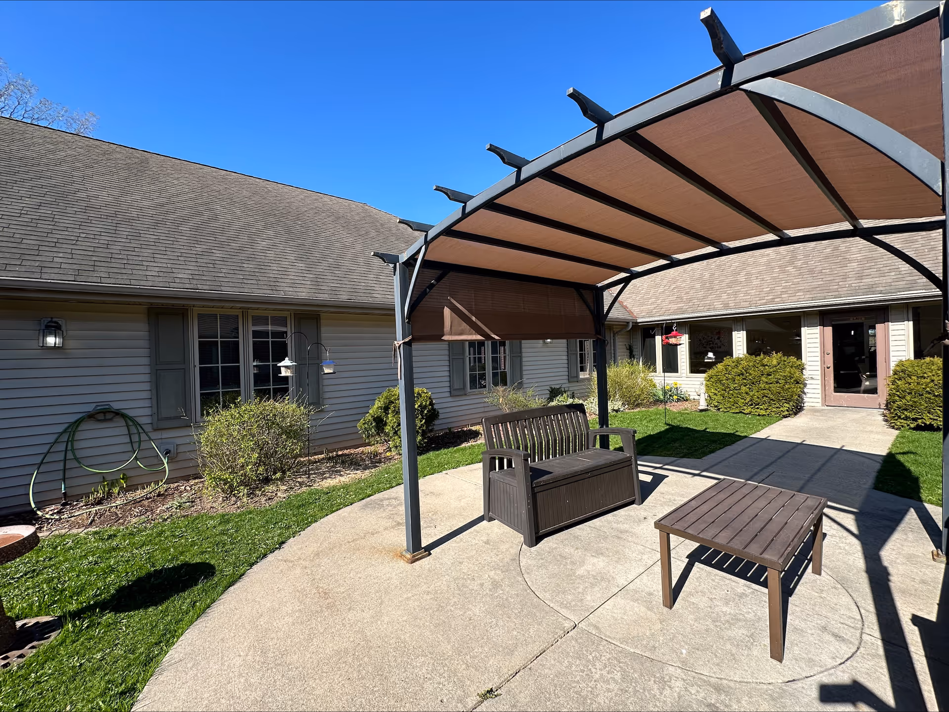 Sunny courtyard with a shaded pergola covering a bench and table in front of a single-story memory care building.